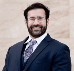 Jacob Scholl, abogado auxiliar en Law Office of Bryan Fagan PLLC, vestido con traje oscuro y corbata, sonriendo frente a un fondo neutro.