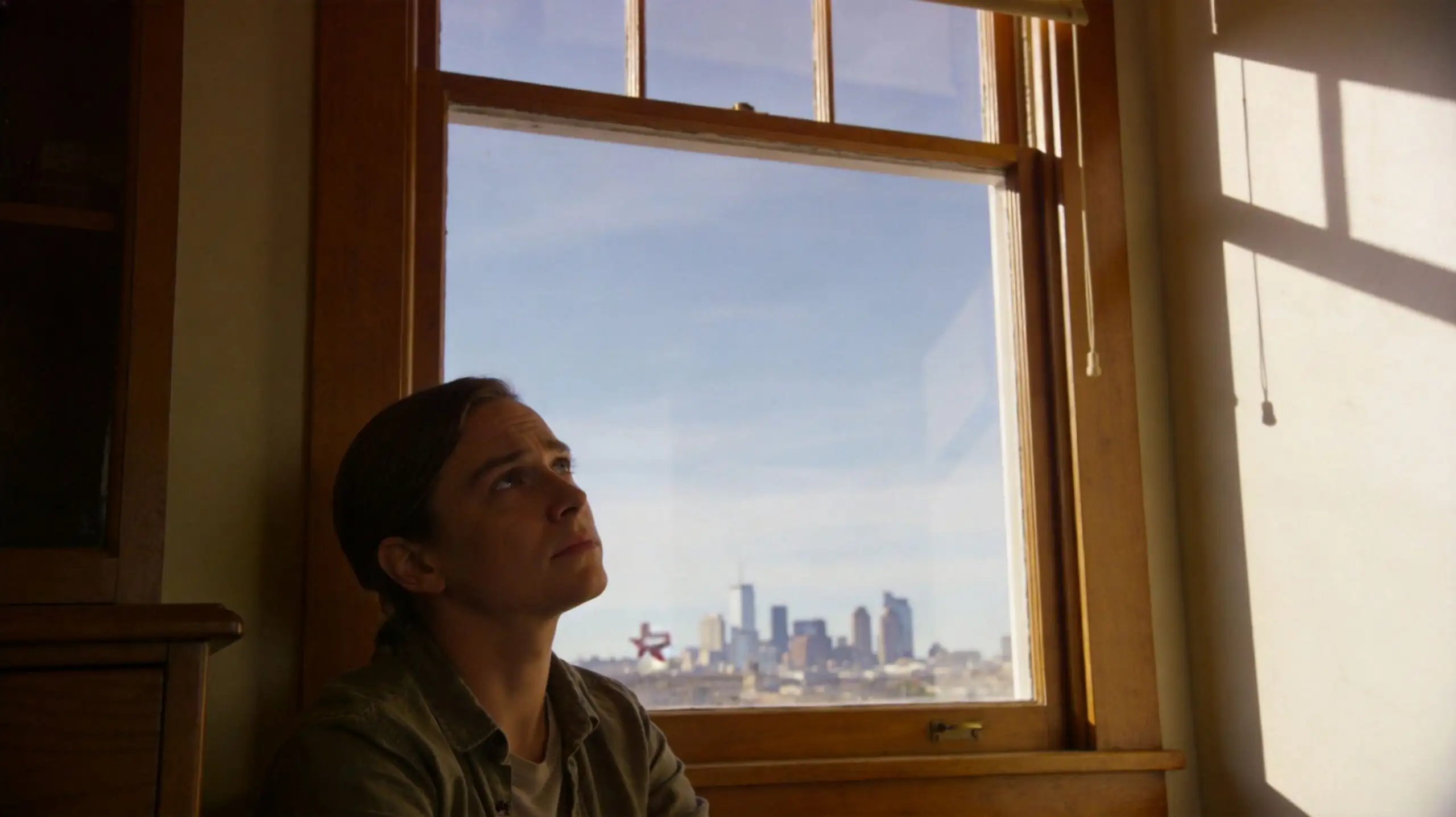 Una persona joven mirando por una ventana con una vista urbana de fondo y luz natural.
